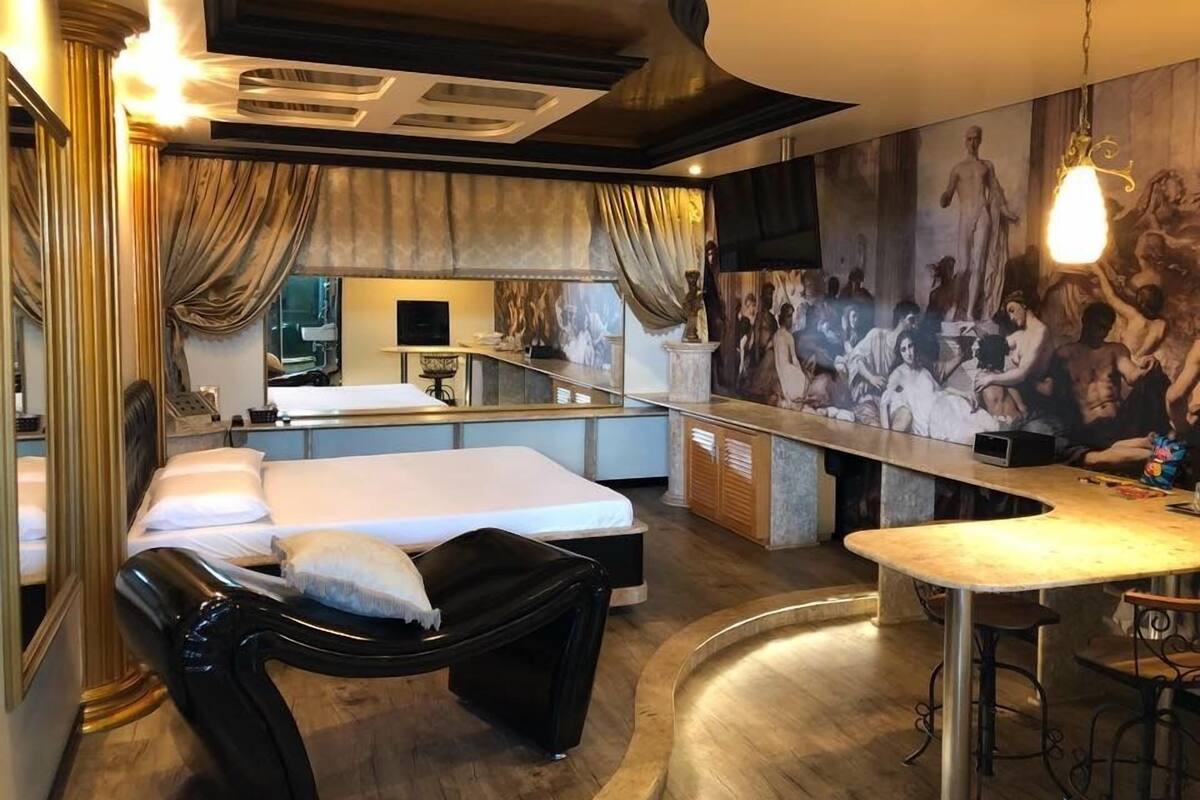 A modern hotel room with a bed, a black chaise lounge, a small bar area, and a mural on the wall.