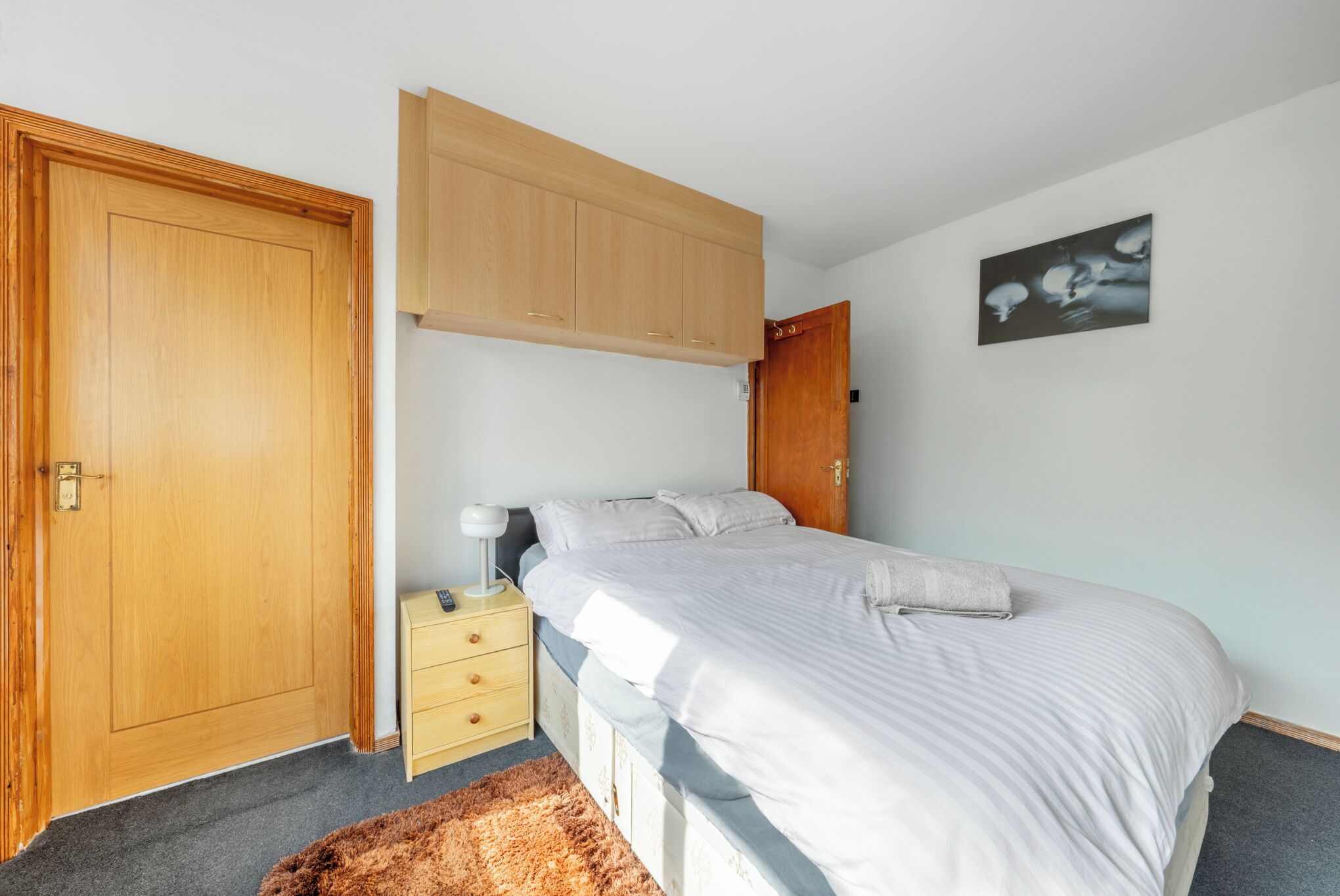 A bedroom with a bed, a nightstand, a wooden wardrobe, a door, and a wall-mounted artwork.