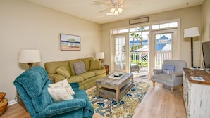 A living room with a green sofa, a blue armchair, a wooden coffee table, and a striped armchair. There is a ceiling fan, a painting on the wall, and a view through the glass doors.