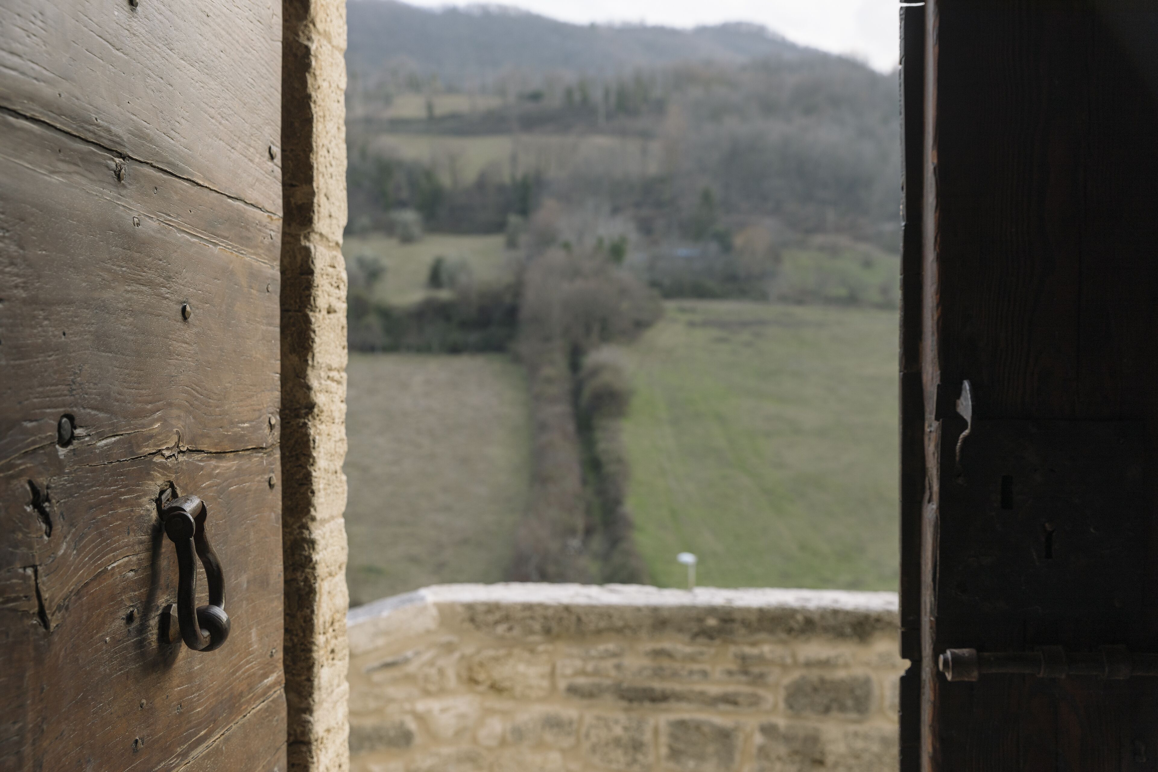 Una puerta de madera rústica con manija de metal, que da a un muro de piedra y una vista de un valle verde con árboles y colinas al fondo.
