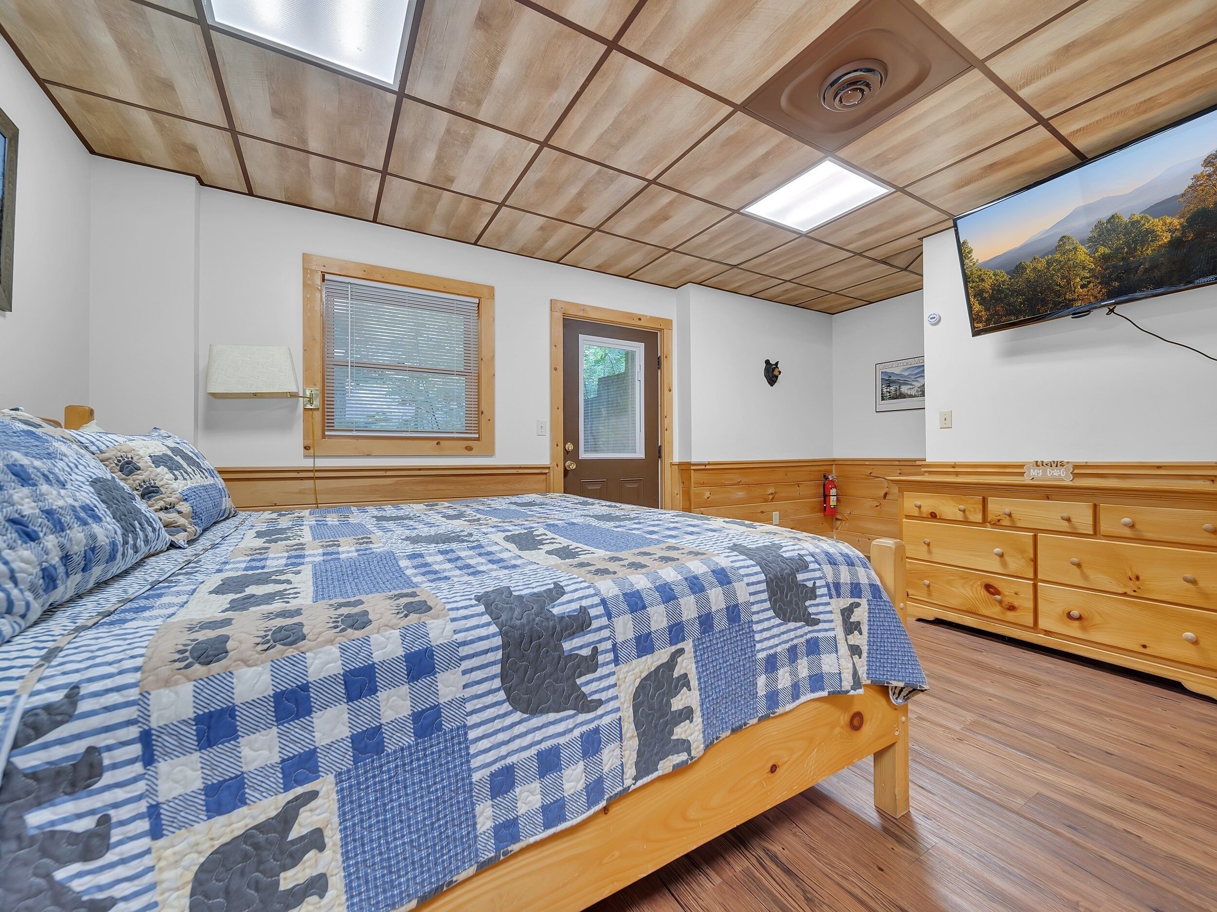 Ein Schlafzimmer mit einem hölzernen Bett, einer Holzkommode, einem Fenster mit Jalousien, einer Tür und einem an der Wand befestigten Fernseher.