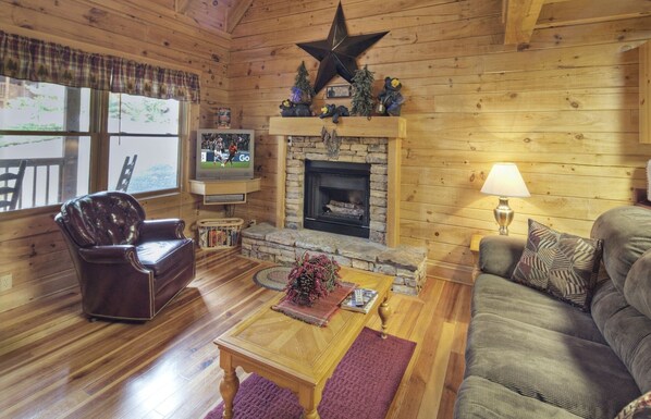 Interior acogedor de una cabaña con chimenea de piedra, paredes de madera, un sillón de cuero, un sofá y un televisor.