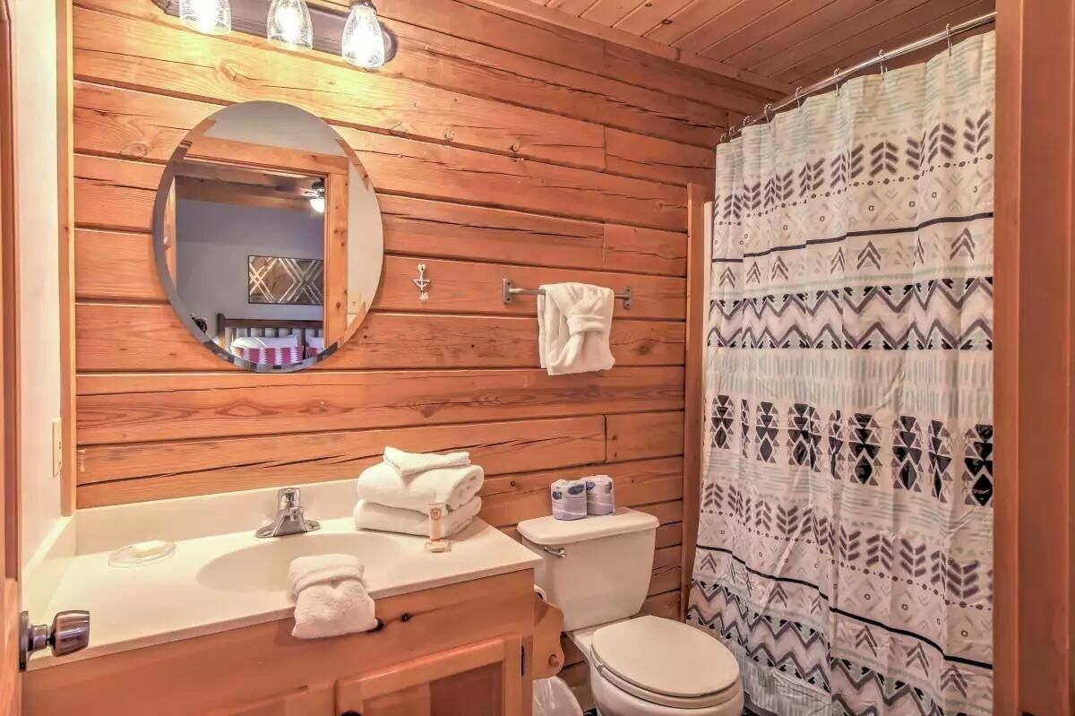 Baño de cabaña de madera con lavamanos, espejo, inodoro y cortina de ducha estampada.