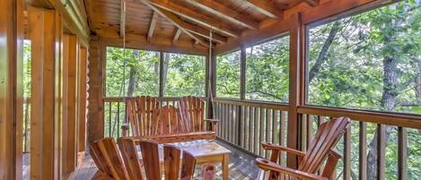 Un patio di legno con poltrone Adirondack e un tavolo, circondato da ampie finestre con vista sulla foresta.