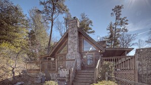 A log cabin with a stone chimney, wooden deck, and a stone staircase leading to the entrance.