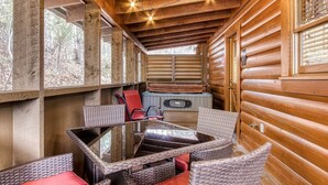 A cozy wooden cabin with a glass-top dining table and wicker chairs, a hot tub, and a window with blinds.