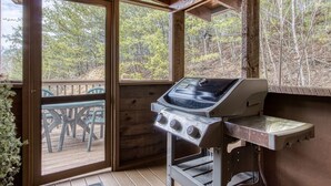A covered outdoor area with a barbecue grill, wooden deck, and a view of the surrounding forest.