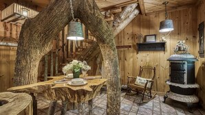 A rustic wooden interior with a dining table set under a large tree trunk, a rocking chair, and a vintage stove.