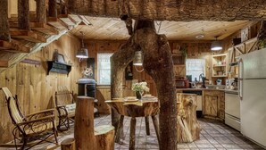 A rustic cabin interior with a wooden staircase, a dining table surrounded by tree stumps, and a kitchen area with modern appliances.