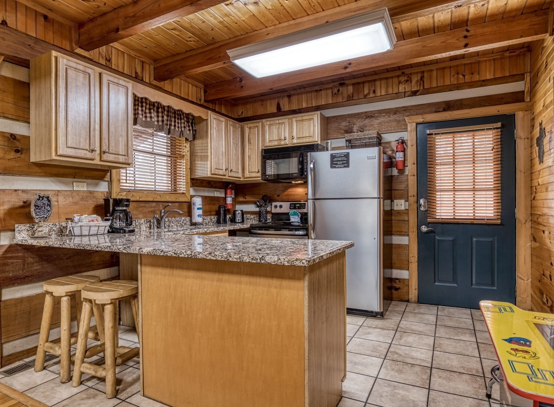 A wooden cabin kitchen with granite countertops, wooden stools, a refrigerator, a microwave, and a door with blinds.