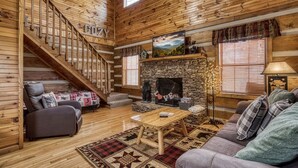 A cozy cabin interior with a stone fireplace, wooden staircase, and a comfortable seating area.
