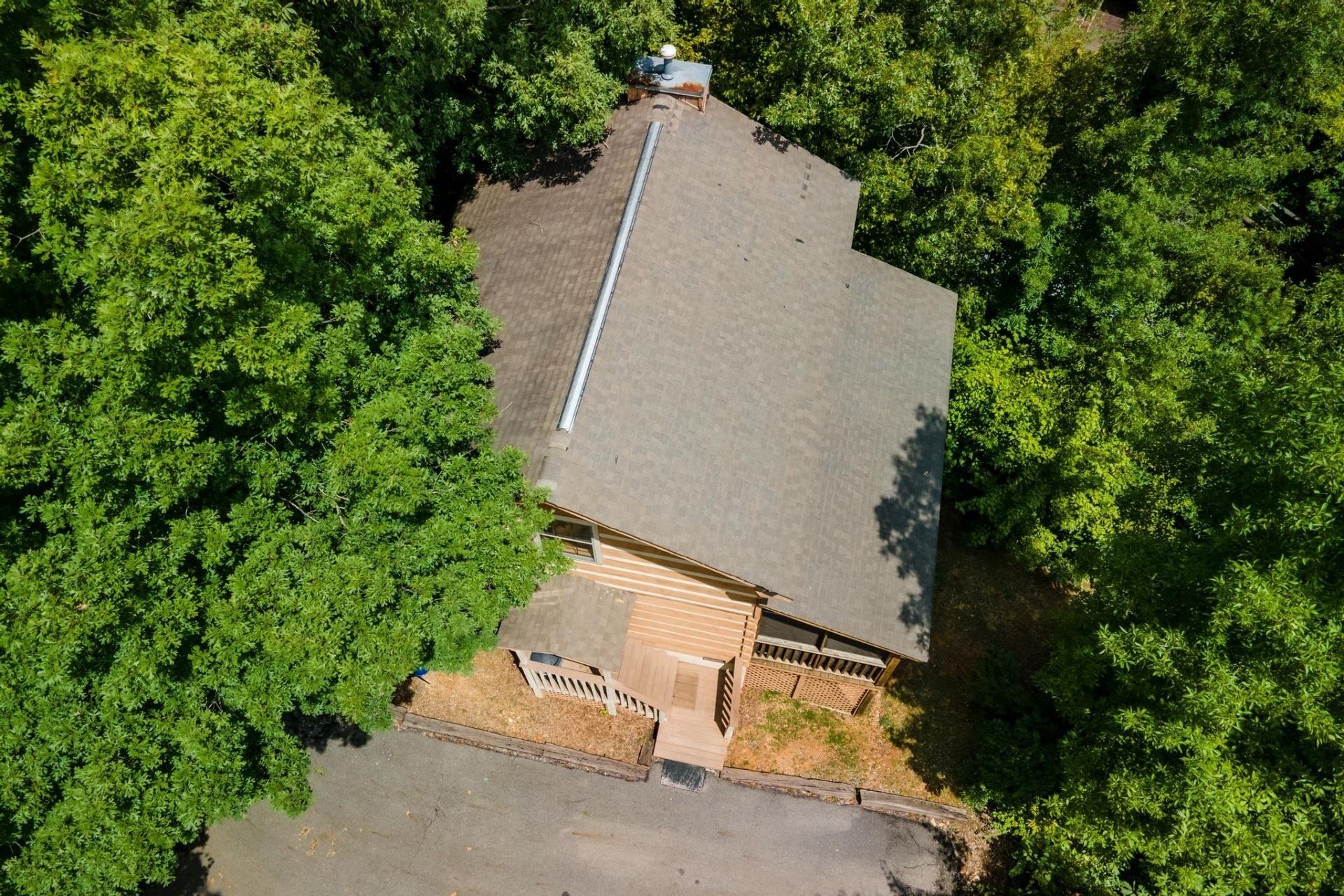 An aerial view of a house surrounded by dense green trees, featuring a sloped roof, a visible chimney, and a small porch area.