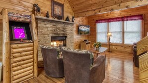 A cozy cabin interior with a stone fireplace, wooden walls, and a television mounted above it. There are two chairs with floral upholstery and a small table with a lamp.