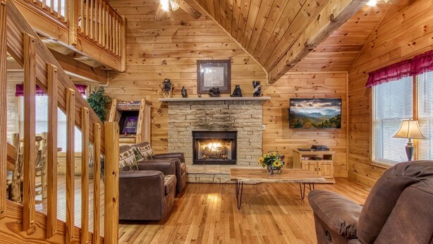 A cozy cabin interior with a stone fireplace, wooden walls, a ceiling fan, and a large window with blinds.