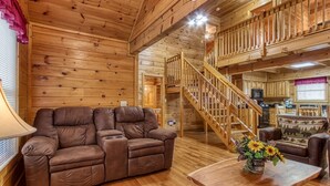 A cozy cabin interior with a wooden interior, a sofa, a table with a vase of flowers, and a staircase leading to an upper level.