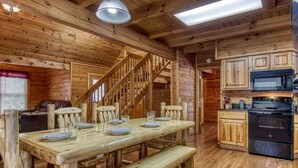 A wooden cabin interior with a dining table, benches, a staircase, a kitchen area with appliances, and a window with blinds.