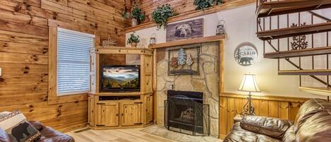 Un interior de cabaña acogedor con chimenea de piedra, paredes de madera, televisor y mobiliario de cuero.