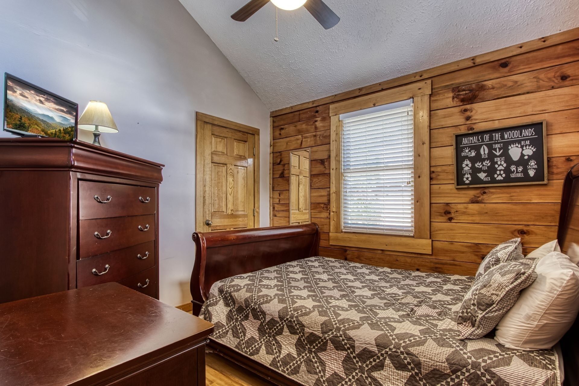 A bedroom with a wooden wall, a bed with patterned bedding, a dresser, a lamp, and a ceiling fan.