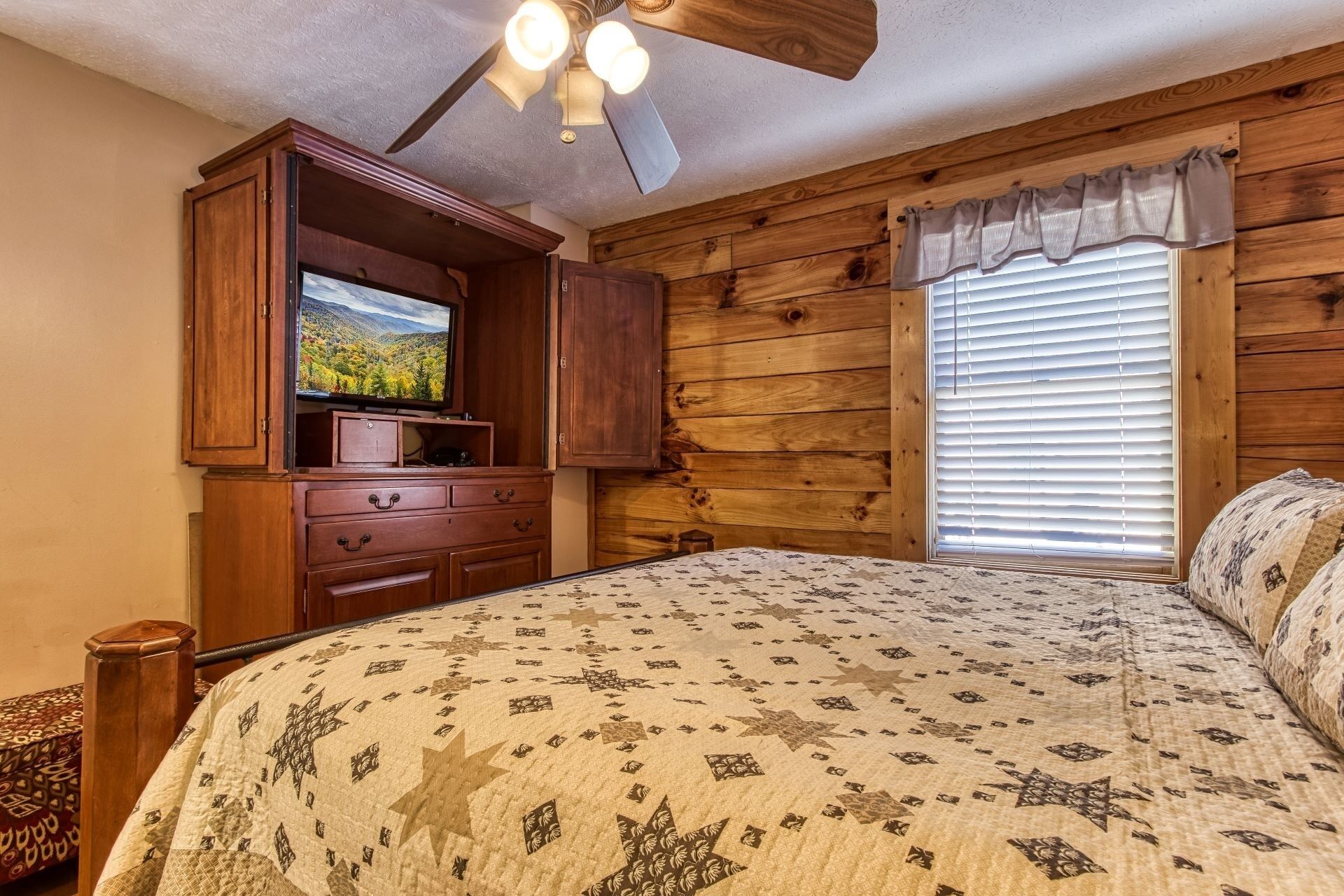 A bedroom with a wooden ceiling fan, a TV on a wooden cabinet, a large bed with a patterned quilt, and a window with blinds.