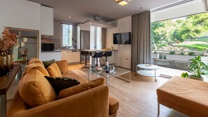 A modern living room with a sofa, coffee table, and a dining area with chairs. There is a kitchen in the background and a large window with a view of greenery. - Phaedrus Living Luxury Flat Vouliagmeni (Vari-Voula-Vouliagmeni)