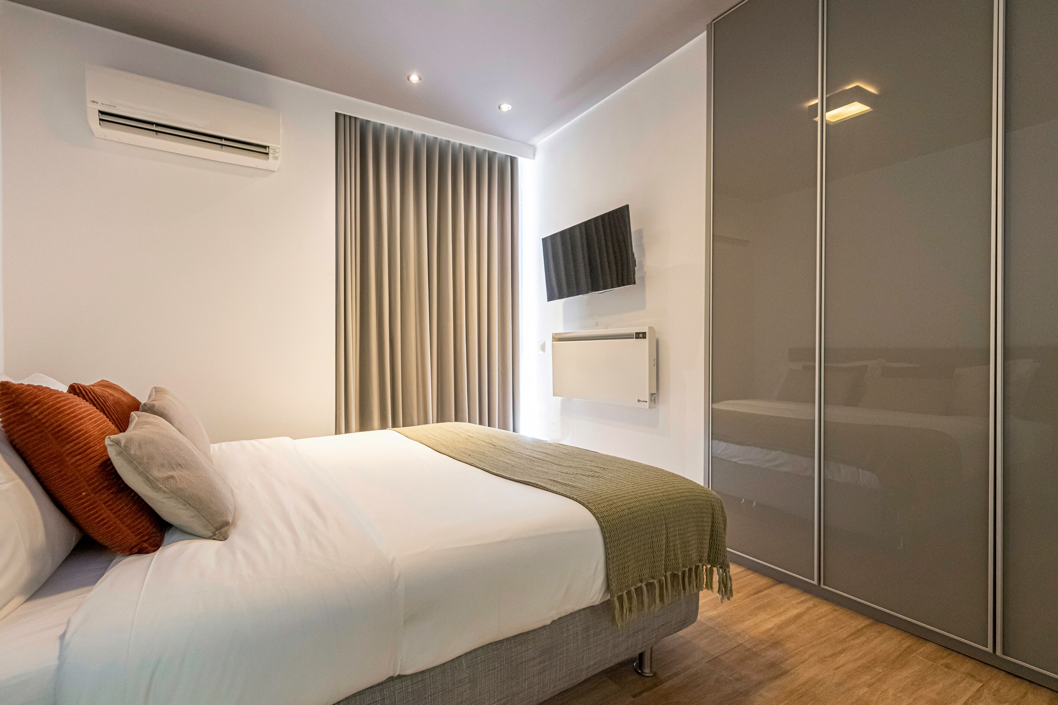 A modern bedroom with a bed, air conditioning unit, window with blinds, TV mounted on the wall, and a wardrobe.