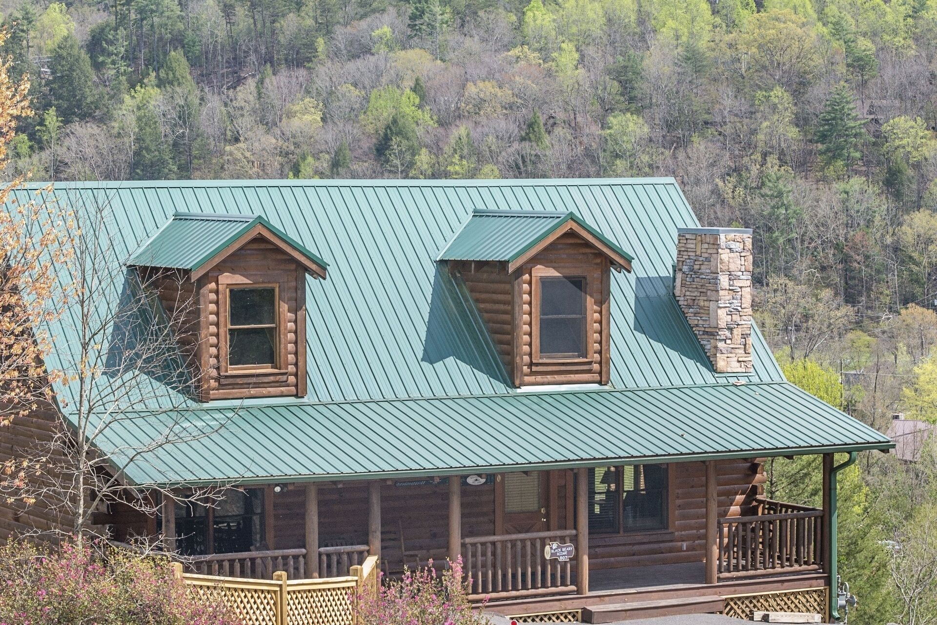 A log cabin with a green metal roof, two dormer windows, and a stone chimney.