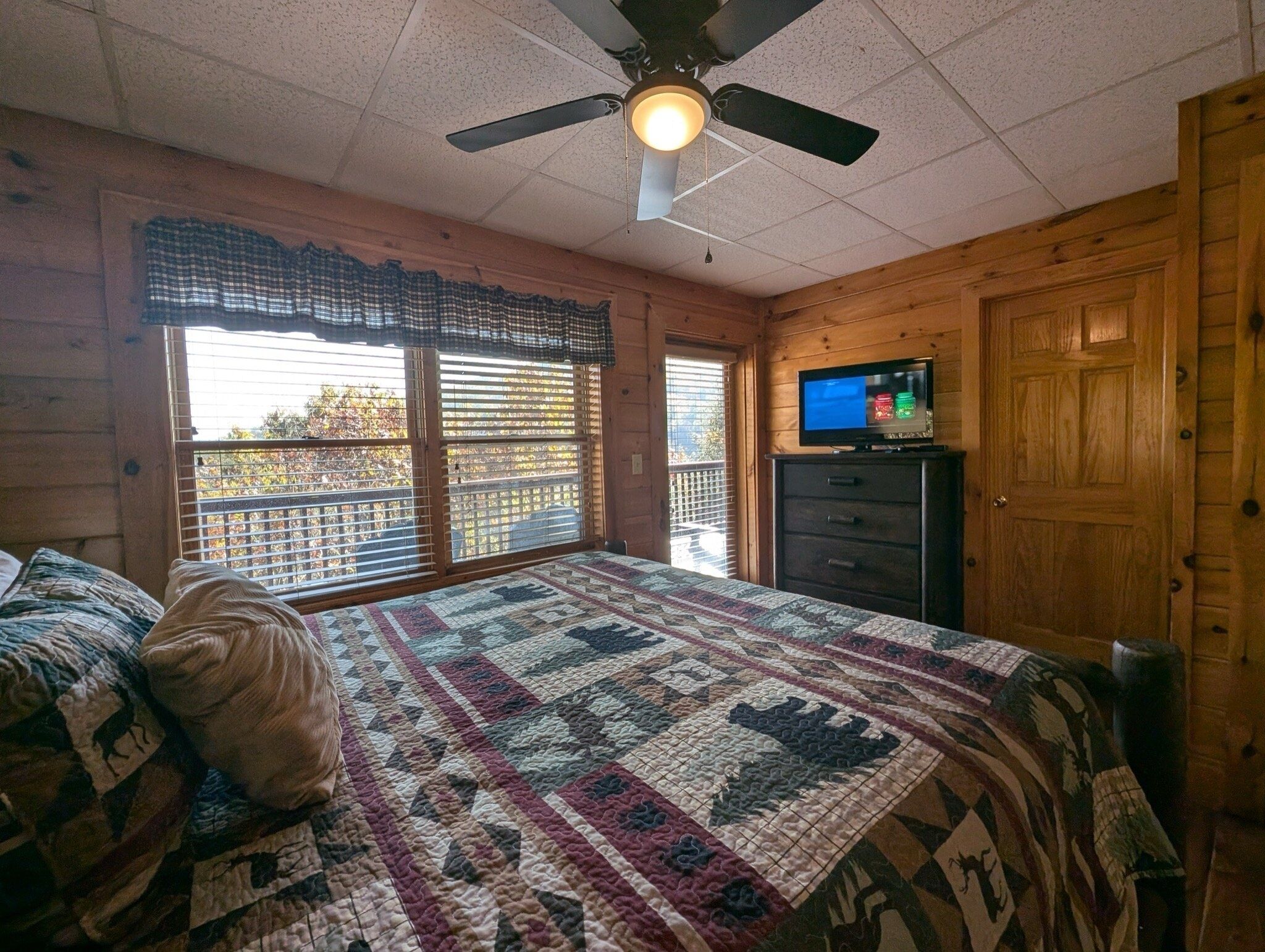 A bedroom with a large bed, a ceiling fan, a television on a dresser, and a wooden door.