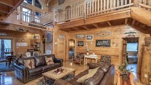 A spacious log cabin interior with a high ceiling, a balcony, and a cozy seating area with leather sofas and a coffee table. - Ern854 - Wagon Wheel Lodge - Great Location! Close To All The Action! 9 Bedroom Cabin by RedAwning (Pigeon Forge)