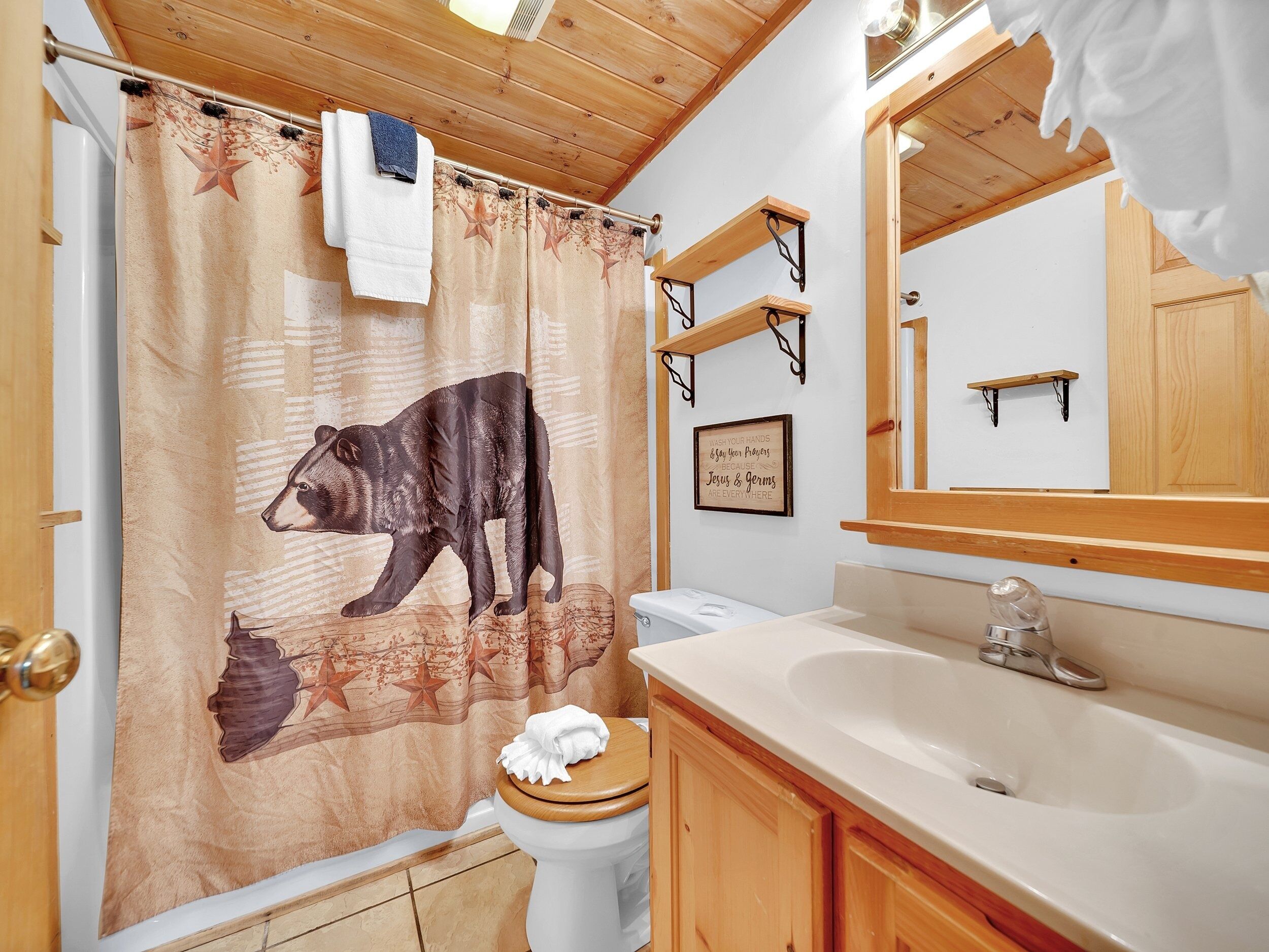 Une salle de bain avec un plafond en bois, un rideau de douche orné d’un motif d’ours, des toilettes, un lavabo et un miroir.