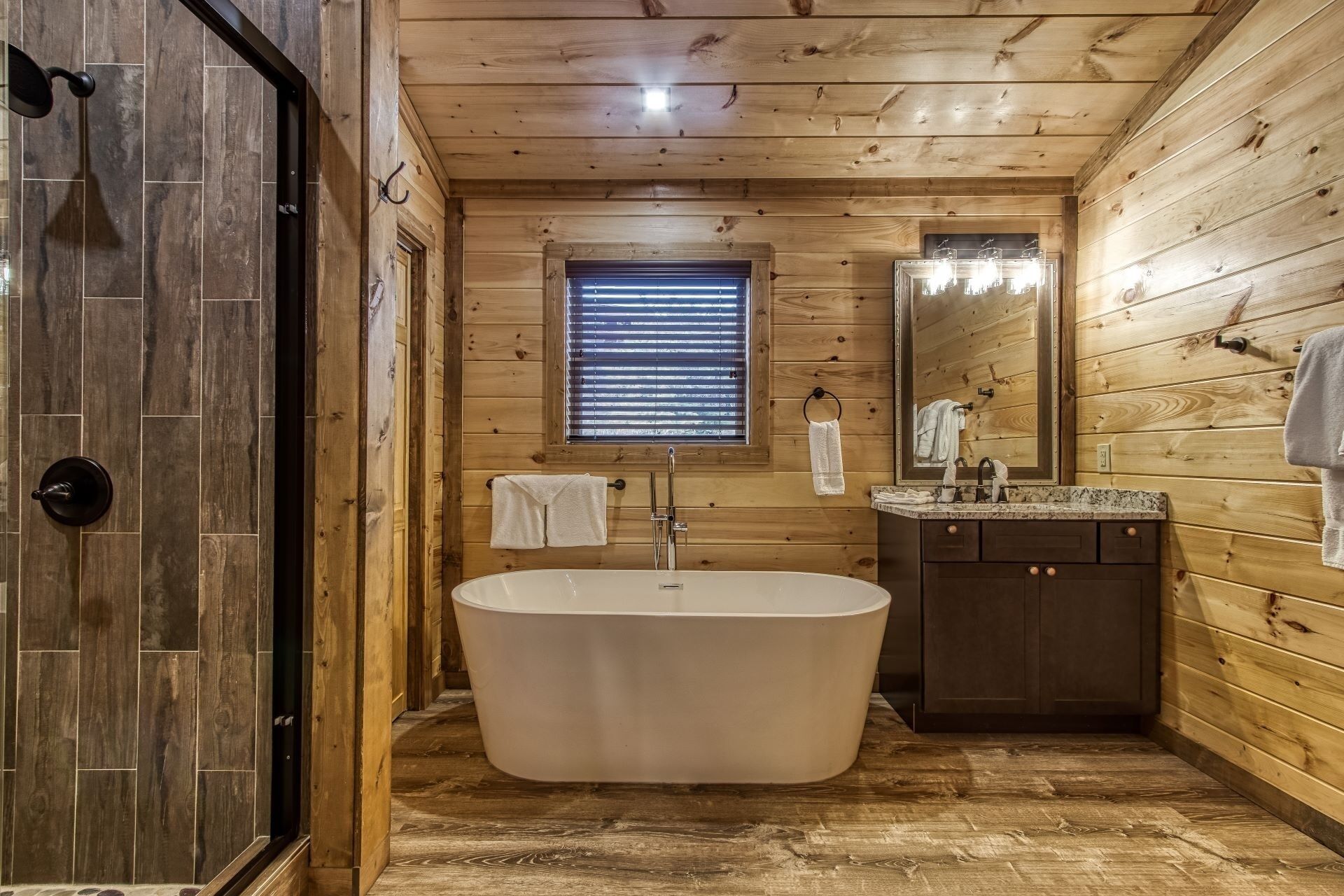 A wooden cabin bathroom with a freestanding bathtub, a vanity with a mirror, and a window with blinds.