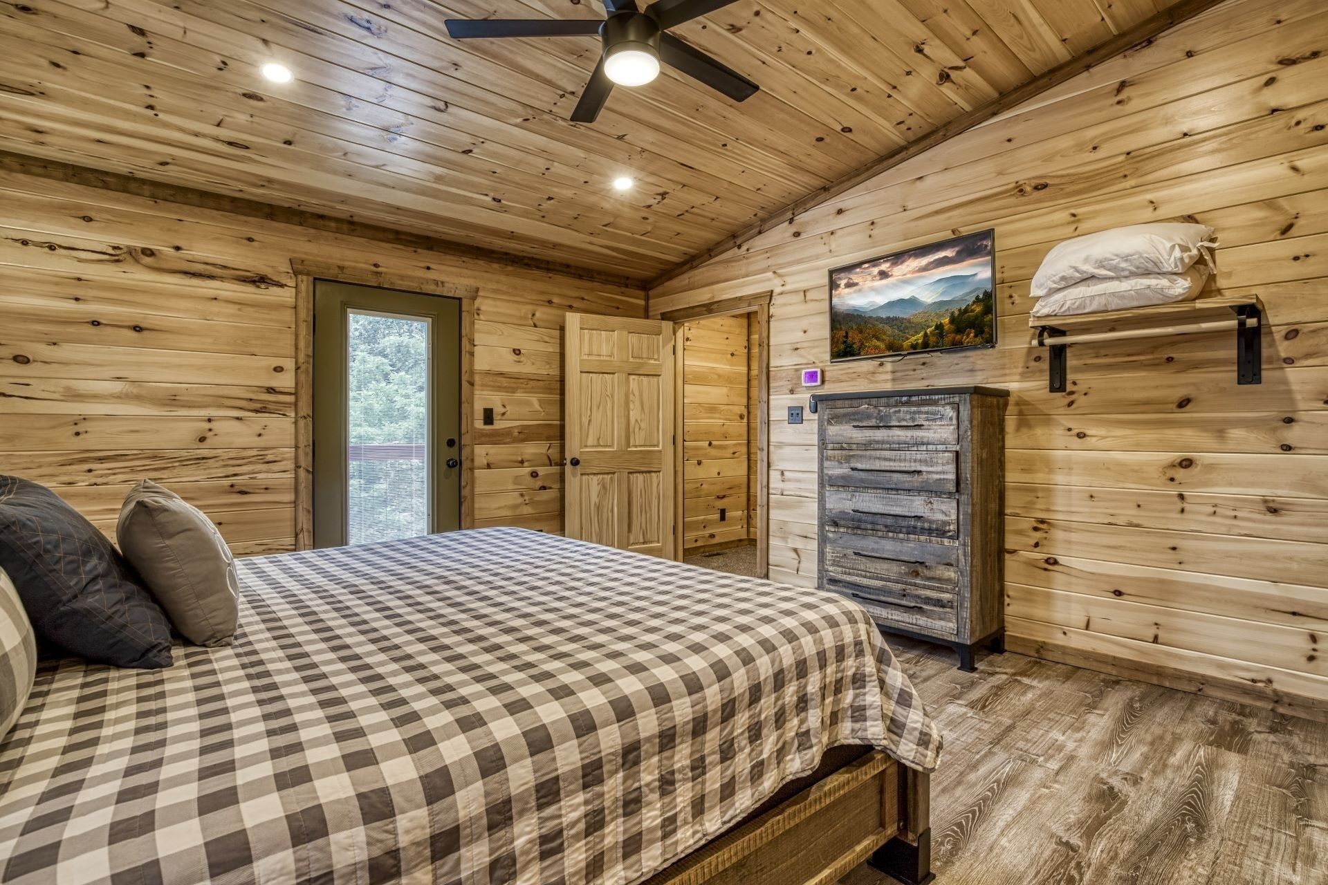 A wooden cabin bedroom with a bed, a refrigerator, and a door leading outside.