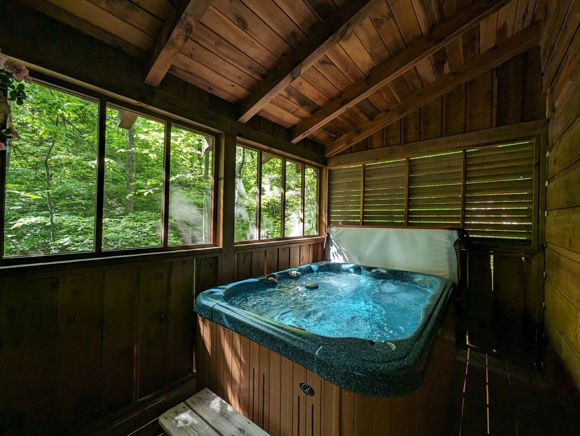 A wooden cabin with a hot tub, large windows, and a view of the forest.