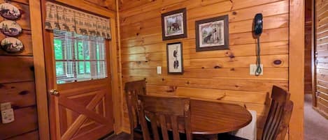 Un interior de madera con una mesa y sillas para comer, cuadros en la pared, un teléfono y una puerta con ventanal.