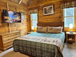 Una camera in una baita di legno con un letto, una cassettiera, una televisione e un quadro appeso al muro.