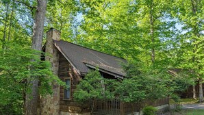 A rustic cabin with a stone foundation, a wooden deck, and a surrounding forest.