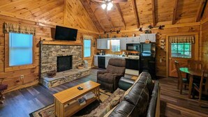 A cozy cabin interior with a stone fireplace, wooden ceiling beams, a television, a kitchen area, and a leather sofa.