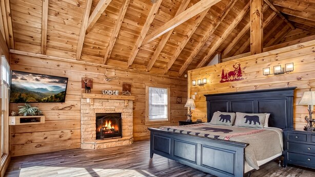 A log cabin bedroom with a bed, a fireplace, a mounted TV, and a window.