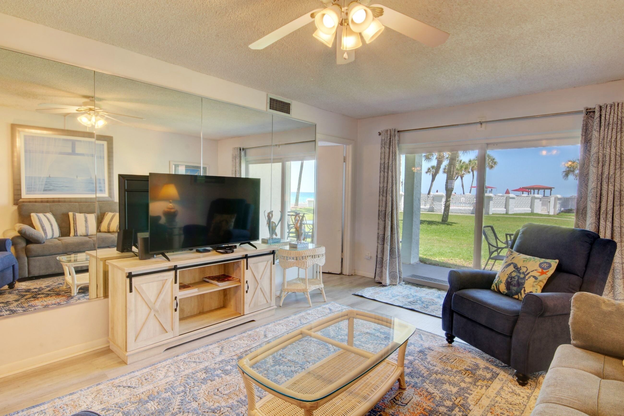 A living room with a television, sofa, and armchair, a large window with a view of the beach, and a ceiling fan.