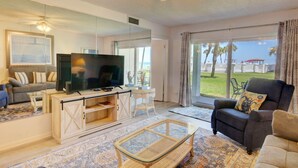 A living room with a television, sofa, and armchair, a large window with a view of the beach, and a ceiling fan.