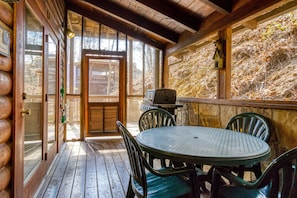 A log cabin with a wooden deck, a round table with chairs, a grill, and a view of a forested hillside.