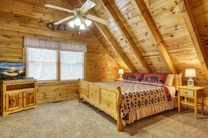 A wooden cabin bedroom with a sloped ceiling, a bed, a nightstand, a lamp, a TV, and a window with blinds.