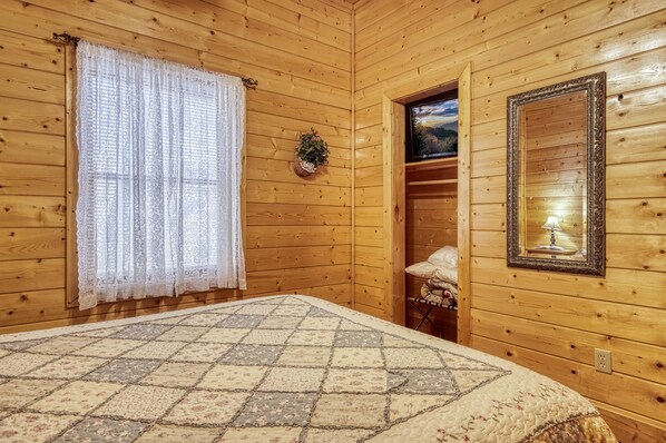 Een houten kamer met een bed, een raam met kantgordijnen, een spiegel en een lijst met een foto aan de muur.