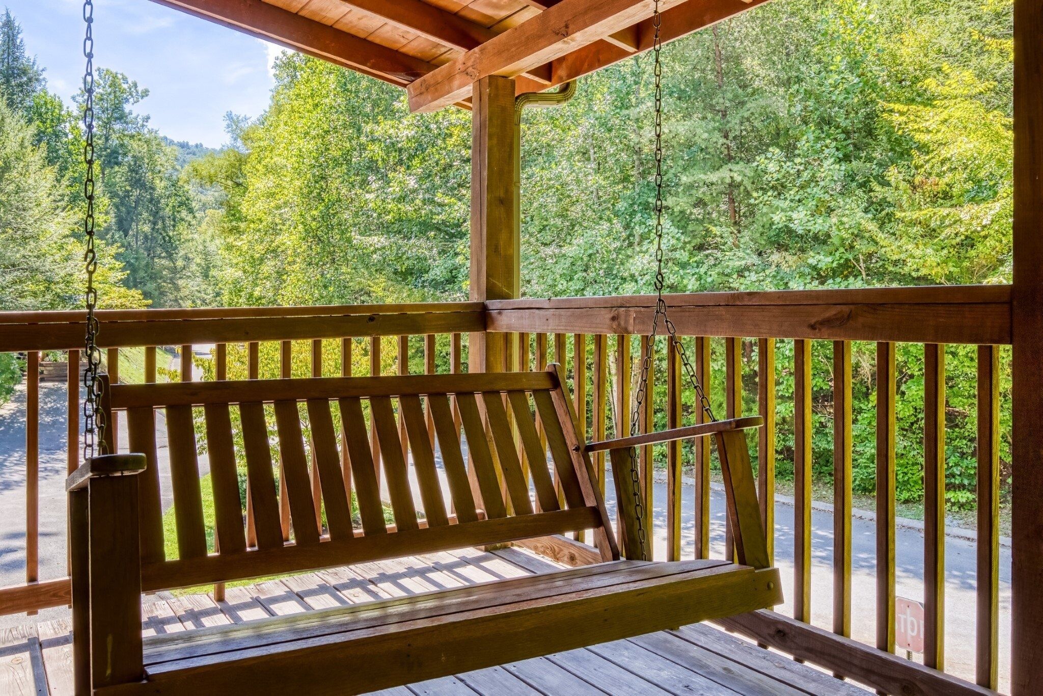 A wooden bench with a metal chain backrest, suspended on a wooden deck with a railing, overlooking a forested area.