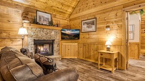 A cozy log cabin interior with a stone fireplace, wooden walls, a comfortable sofa, and a small table with a lamp. - Er 229 Brief Escape - Great Location Close to Town - 4 BR Cabin by RedAwning (Pigeon Forge)