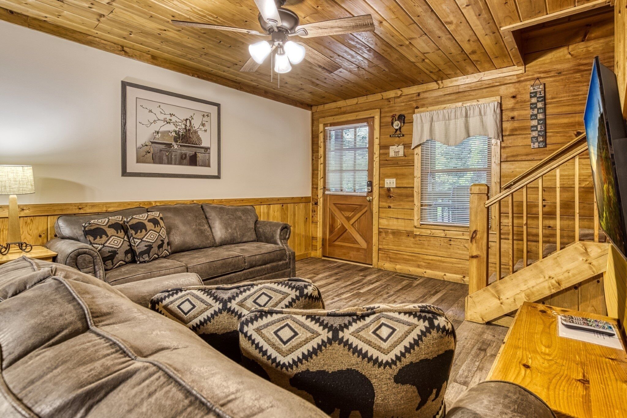 A cozy wooden cabin interior with a sofa, a coffee table, a ceiling fan, and a staircase.