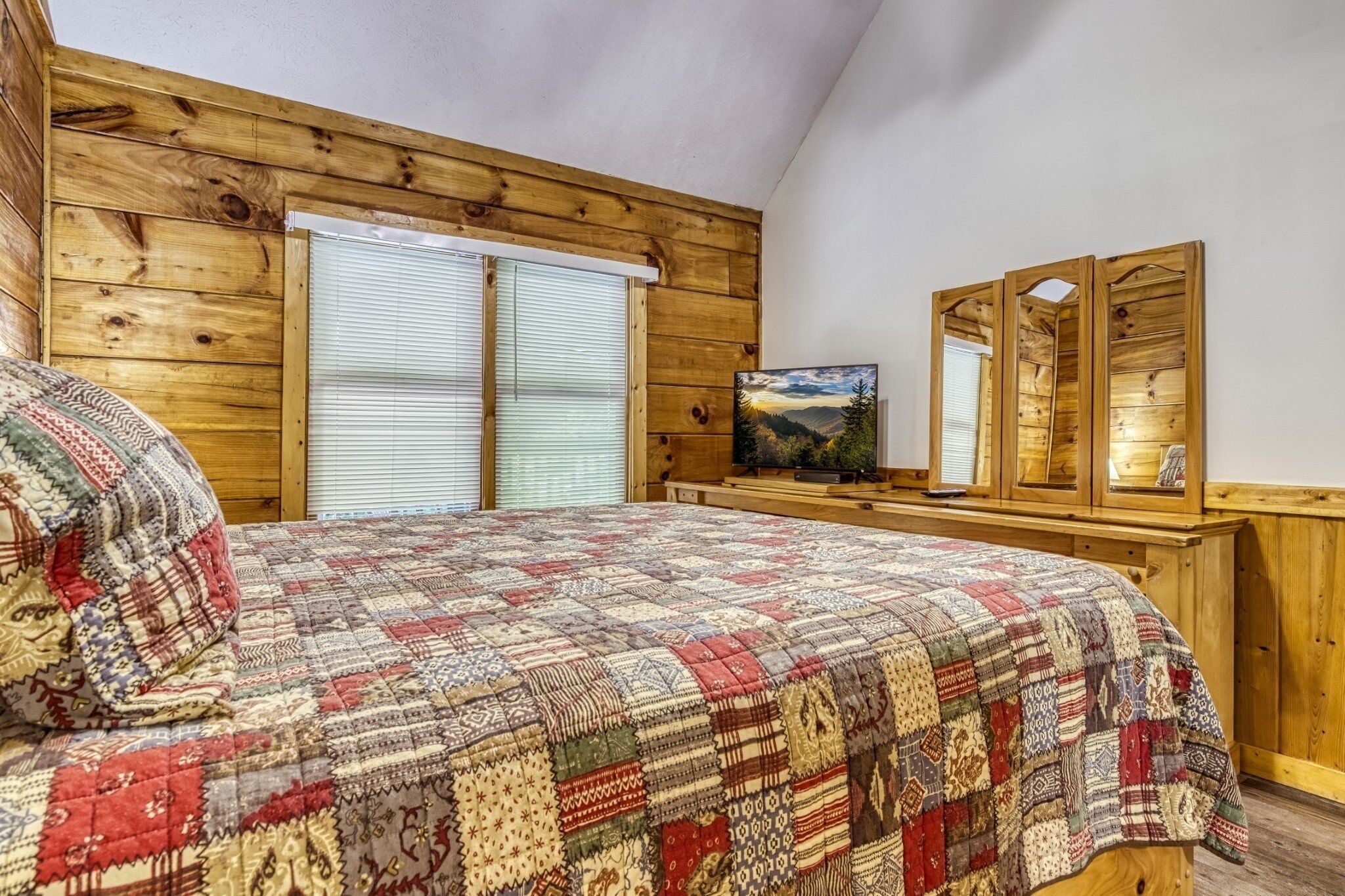A bedroom with a wooden ceiling, a bed with a patterned quilt, a television, and a mirror on a wooden dresser.