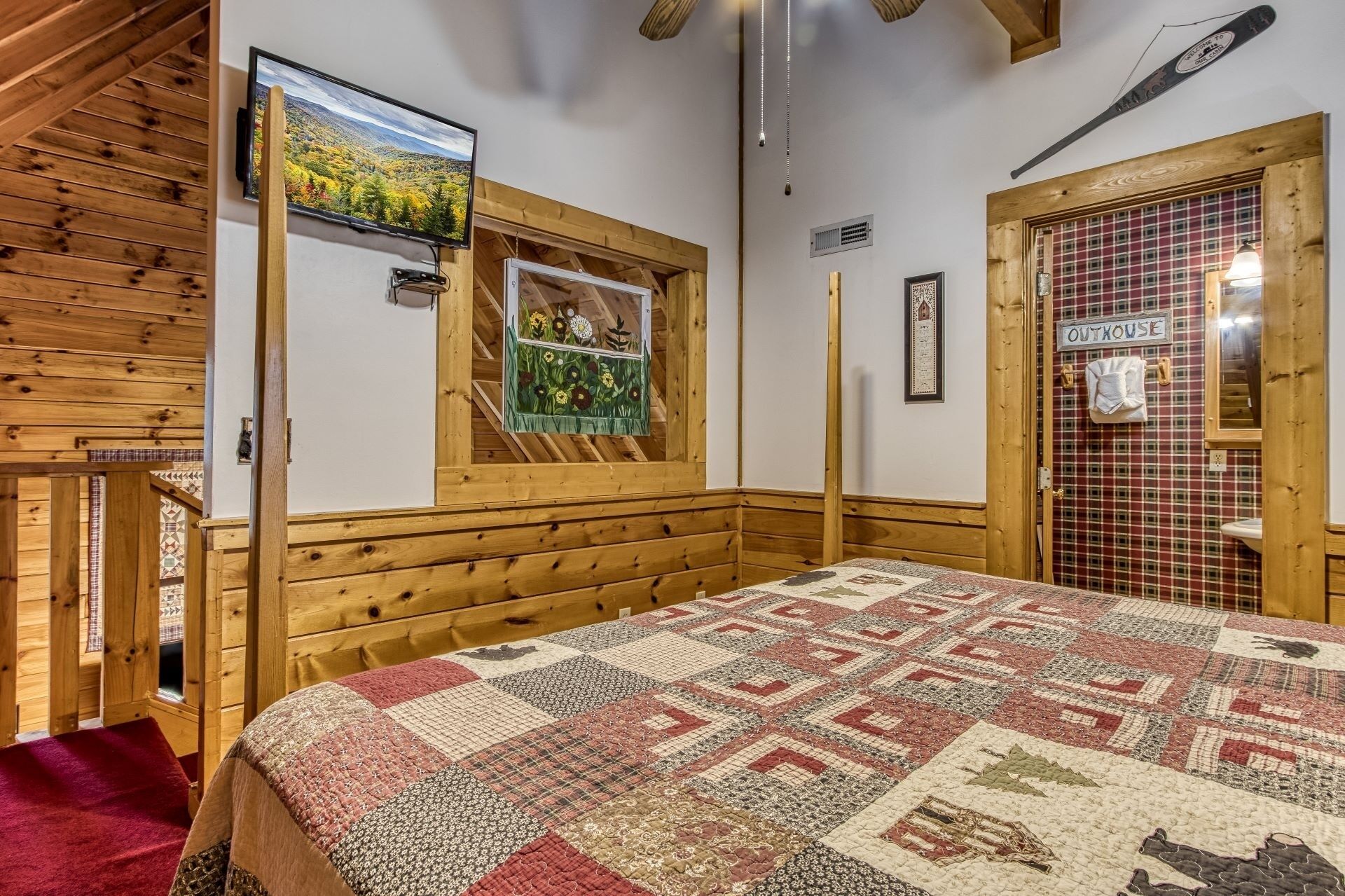 A cozy room with a wooden sloped ceiling, a quilted bedspread, a mounted TV, a window with a view of trees, and a door labeled "OUTKOUSE".