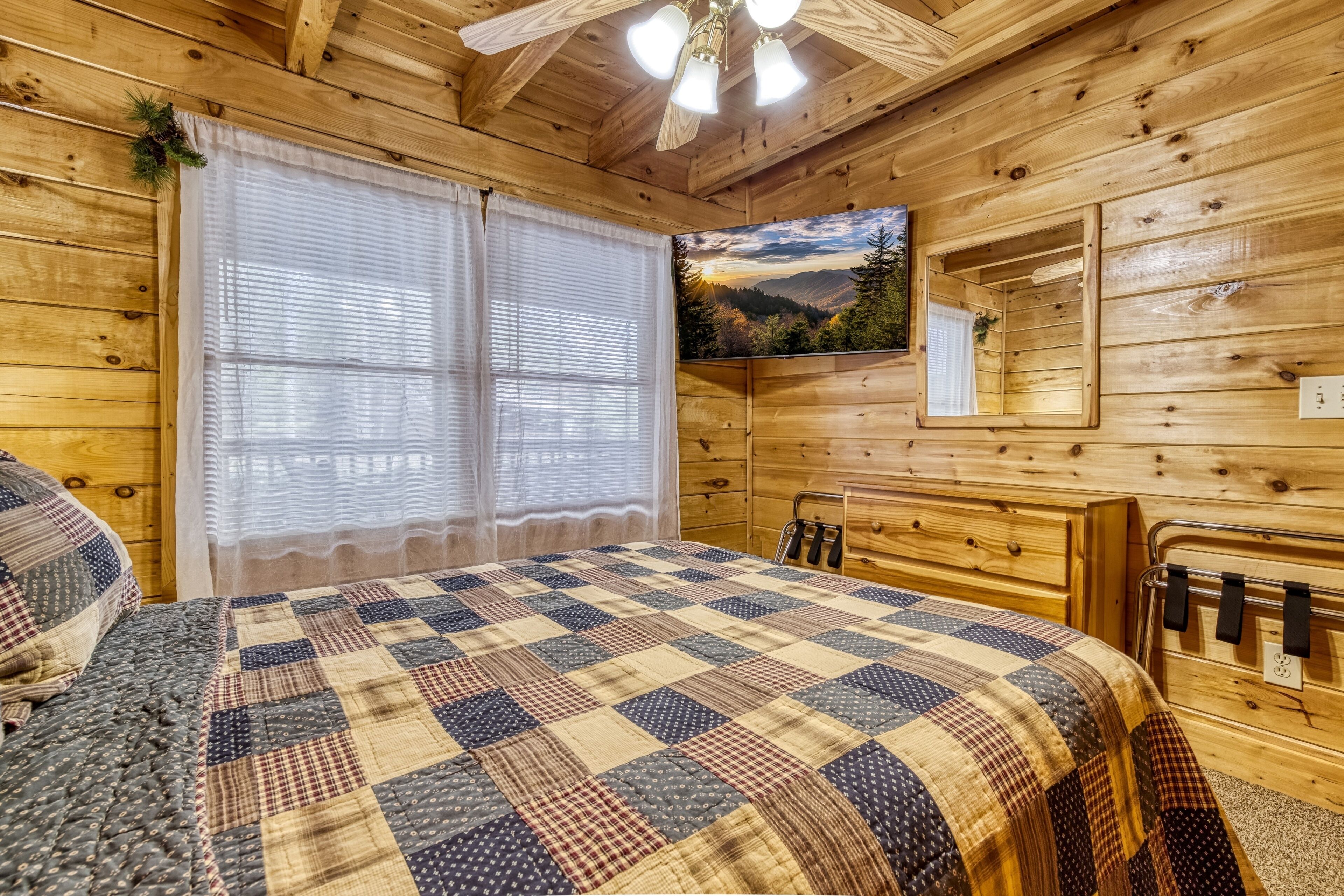 A wooden cabin bedroom with a large bed, a window with curtains, a painting of a landscape, and a wooden dresser with a mirror.