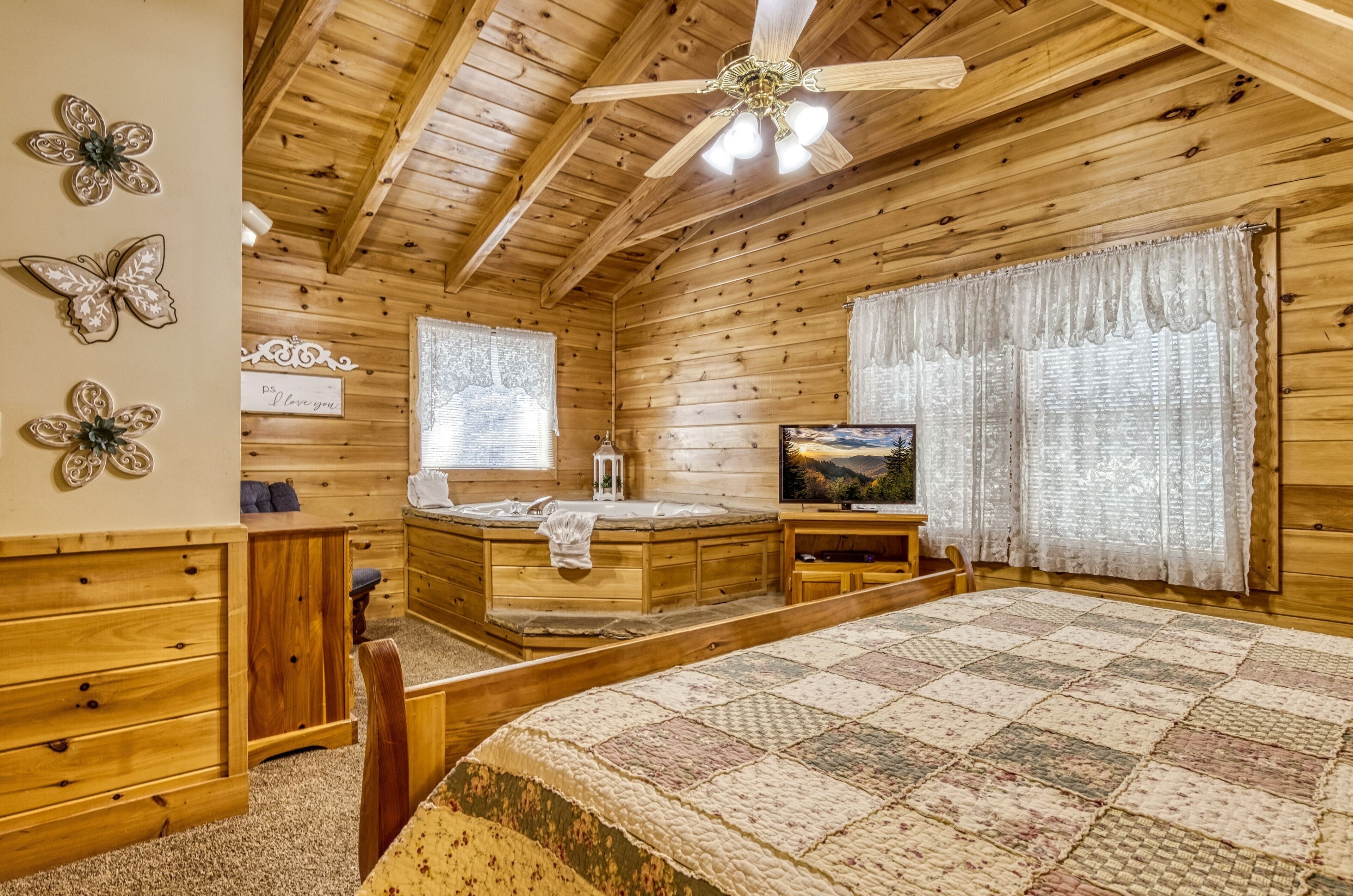 Un dormitorio tipo cabaña de madera con una cama grande, un escritorio de madera, un televisor, una bañera de hidromasaje y mariposas decorativas en la pared.