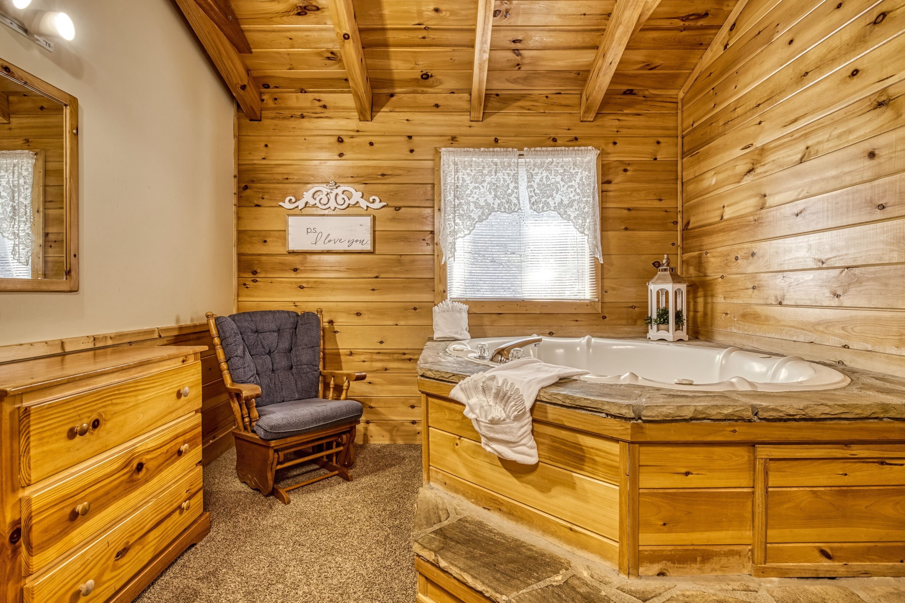 Une salle de bain rustique, avec des murs en bois, comprenant une chaise en bois, un meuble-lavabo, une baignoire et une fenêtre ornée de rideaux en dentelle.
